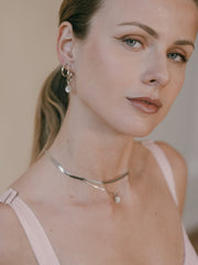 A woman with long hair tied back looks at the camera. She wears a pink top, a sterling silver necklace, and Elsa Peony Sapphire Earrings by CAMELLIA handcrafted in New York.