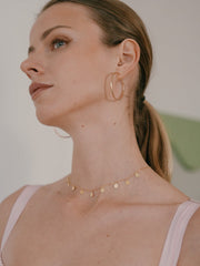 A woman with light brown hair in a ponytail wears large hoop earrings and the CAMELLIA Mother Chain Necklace crafted in 14K Yellow Gold Vermeil, featuring small circular pendants. She is gazing upwards, dressed in a pale pink top.
