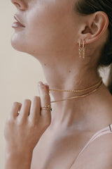 The person flaunts CAMELLIA's Willo Cz Chain Hoop Earrings and a ring with a dark stone, beautifully paired with layered 14K Yellow Gold Vermeil necklaces. They touch the necklaces gracefully, exuding an aura of elegance.
