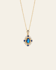 The CAMELLIA Eta Sapphire Necklace is a gold vermeil piece featuring an oval blue sapphire gemstone, elegantly surrounded by smaller blue and clear stones in a decorative pendant.