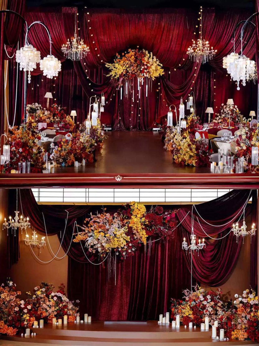 A lavish indoor setting features red drapery, chandeliers, abundant floral arrangements, candles, and a CAMELLIA burgundy Theater Style Wedding Flower Ring masterpiece to create an elegant event atmosphere.