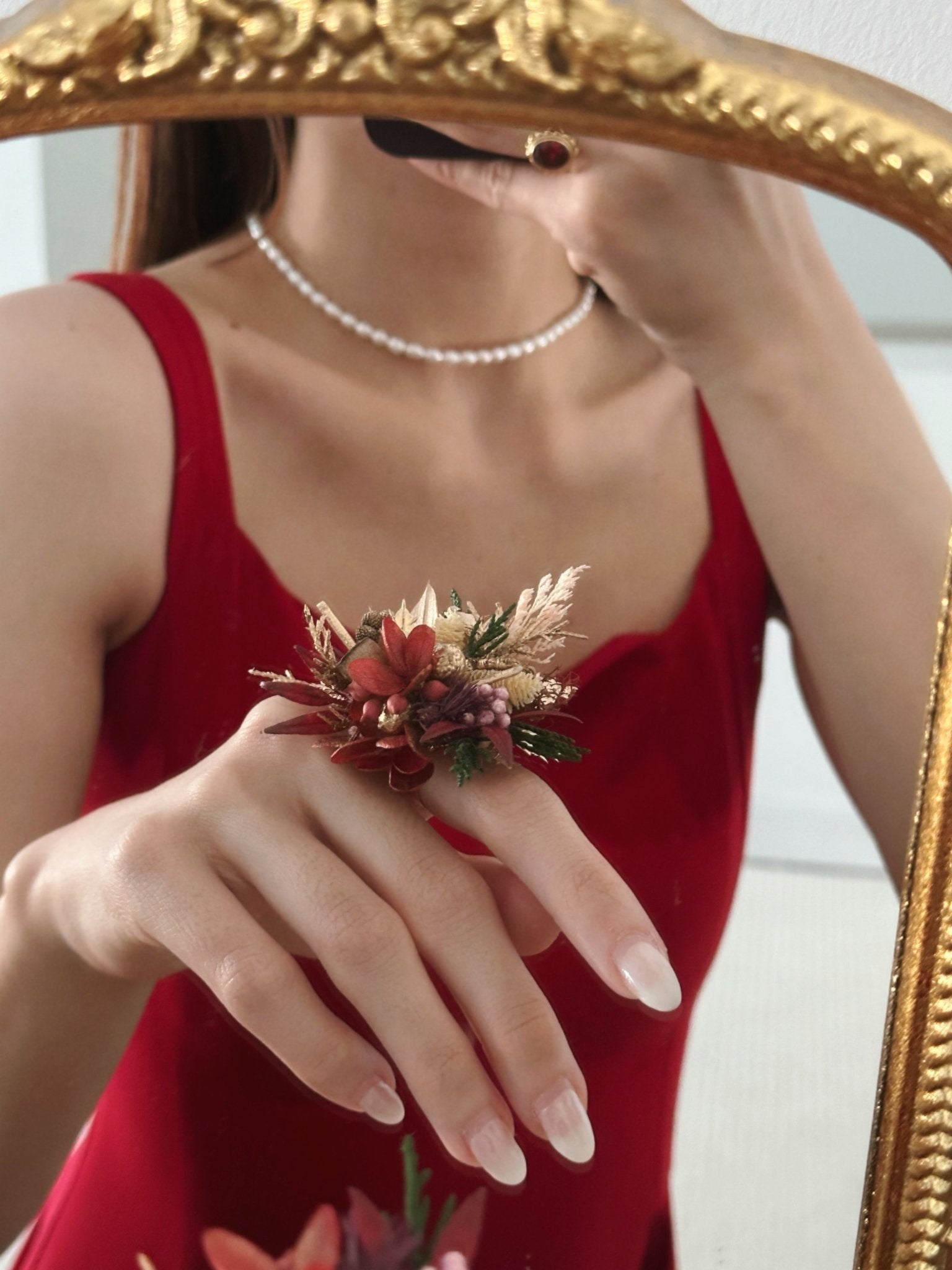 A person in a red dress, wearing a pearl necklace, takes a mirror selfie showcasing CAMELLIA's Customized Wedding Flower Ring on their left hand, featuring striking red and beige flowers.