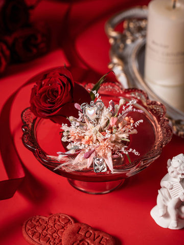 The Confession Valentine’s Day Flower Ring from CAMELLIA features a red rose, "LOVE" heart-shaped cookies, and dried preserved flowers in a glass dish on a red backdrop. Handcrafted in New York, it adds charm to any setting. The Confession Valentine’s Day Flower Ring from CAMELLIA features a red rose, "LOVE" heart-shaped cookies, and dried preserved flowers in a glass dish on a red backdrop. Handcrafted in New York, it adds charm to any setting.