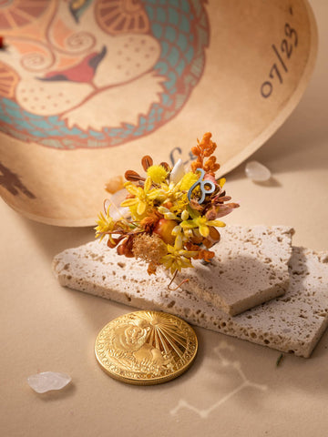 The Leo Flower Ring by CAMELLIA is showcased among a golden coin, a small bouquet of yellow and orange flowers, and stone pieces on a surface, with a decorative painted object partially visible in the background, resembling an exquisite handcrafted treasure from New York. The Leo Flower Ring by CAMELLIA is showcased among a golden coin, a small bouquet of yellow and orange flowers, and stone pieces on a surface, with a decorative painted object partially visible in the background, resembling an exquisite handcrafted treasure from New York.