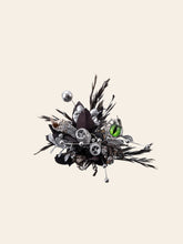 The Devil's Eye Flower Ring by CAMELLIA is a unique handcrafted New York design. Featuring silver gears, black feathers, and a bright green gem reminiscent of abstract floral art, it creates captivating contrast against a plain background.