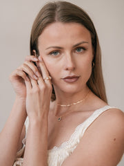 A woman with long light brown hair wearing a white top, gold vermeil hoop earrings, a pavé-set accents necklace, and the CAMELLIA Evermine Moissanite Band poses with her hands near her face against a neutral background.