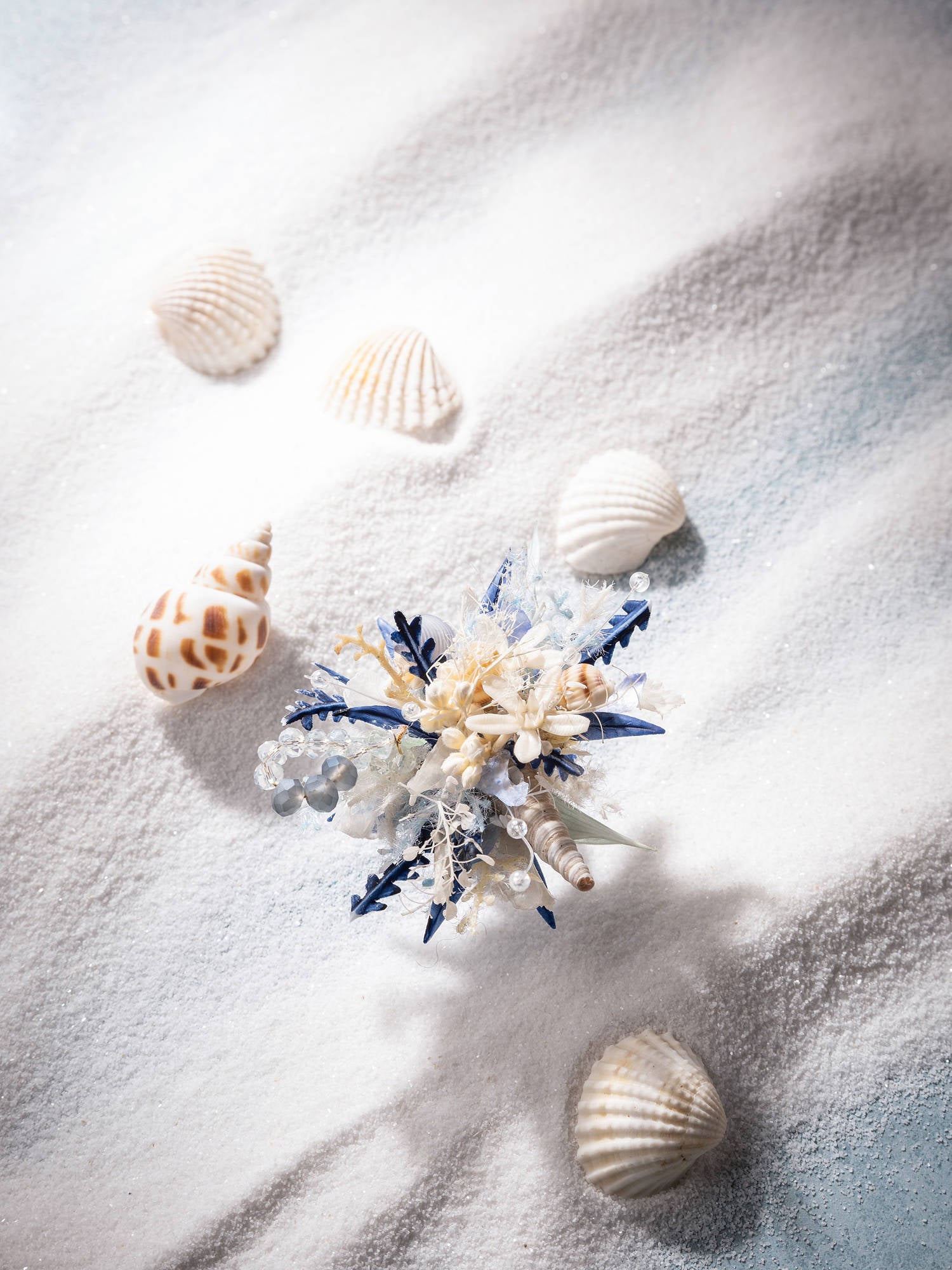 The Ocean Flower Ring by CAMELLIA, a handcrafted decorative piece with blue and white accents from New York, rests on white sand amidst seashells.