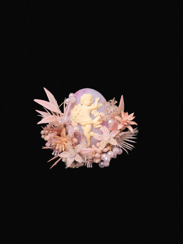 A decorative arrangement with a cherub amidst pastel pink and beige flowers on black, reminiscent of CAMELLIA's Cherry Blossom Birth Flower Ring. A decorative arrangement with a cherub amidst pastel pink and beige flowers on black, reminiscent of CAMELLIA's Cherry Blossom Birth Flower Ring.