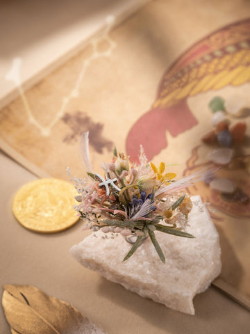 A small bouquet of dried flowers rests on a white rock, accompanied by a gold coin, the CAMELLIA Pisces Flower Ring, a dried leaf, and an open book with illustrations on a beige surface. A small bouquet of dried flowers rests on a white rock, accompanied by a gold coin, the CAMELLIA Pisces Flower Ring, a dried leaf, and an open book with illustrations on a beige surface.