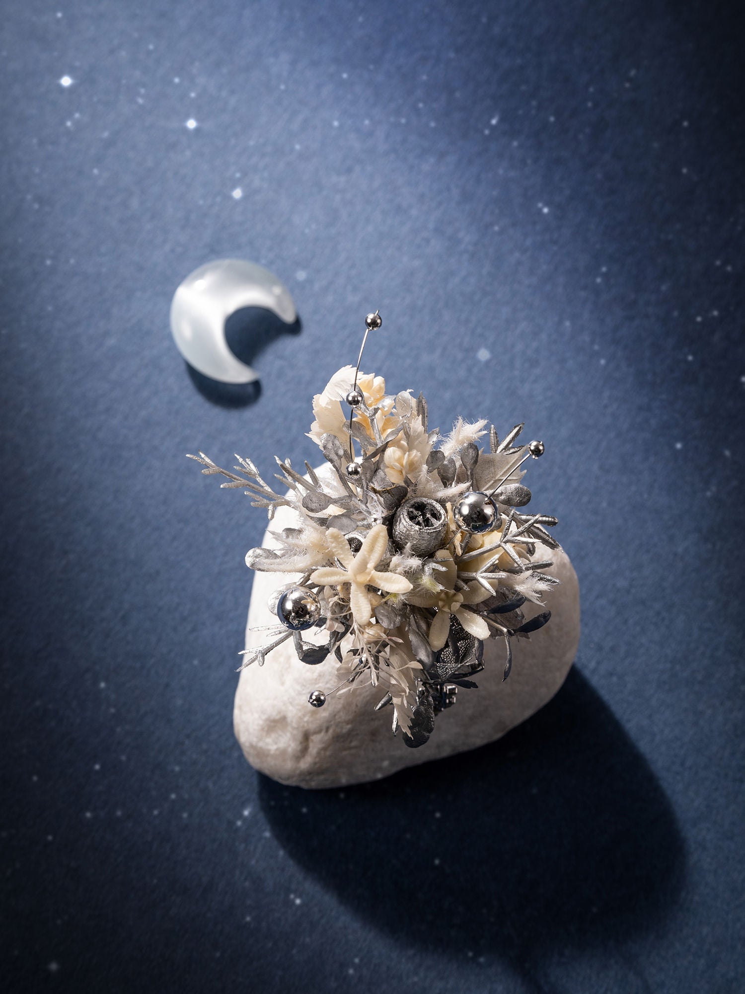 CAMELLIA's Moon Flower Ring, featuring dried preserved flowers and metallic elements on a stone, creates an enchanting display inspired by a crescent moon set against a starry backdrop.