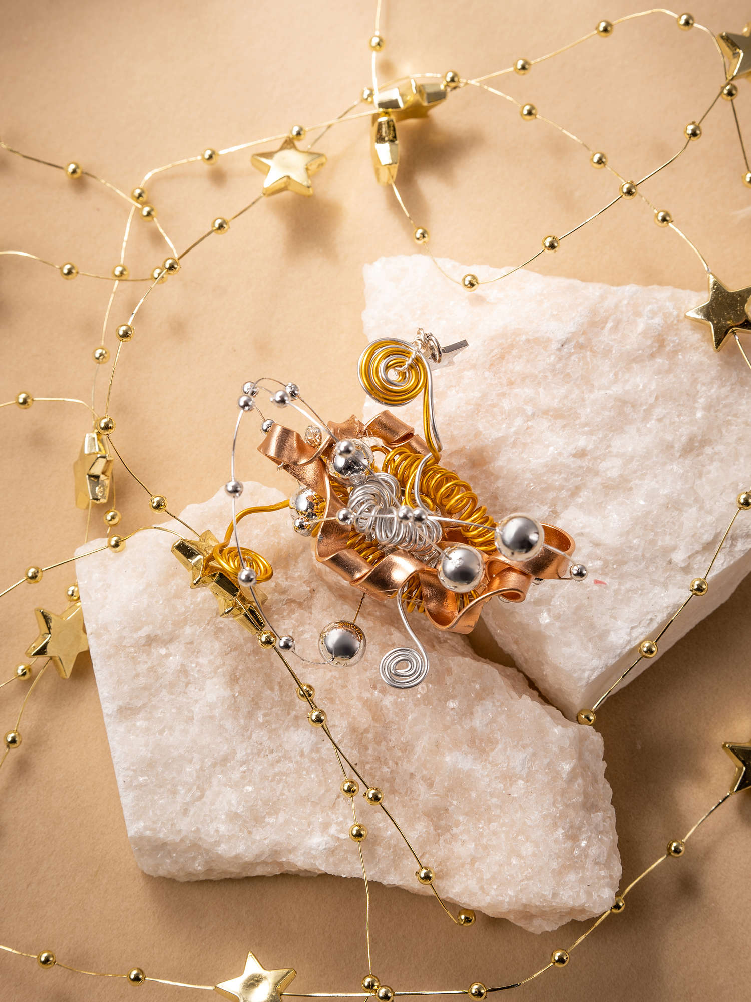 CAMELLIA's Star Flower Ring, paired with festive earrings featuring metallic spirals and beads on white stones, is adorned by a gold star and bead garland on a beige background, creating an elegant handcrafted jewelry ensemble.