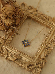 The Cámellia Sapphire Pearl Necklace by CAMELLIA, a gold floral design with sapphire stones, is placed on an ornate 14K Yellow Gold Vermeil picture frame beside dried roses on a beige surface.