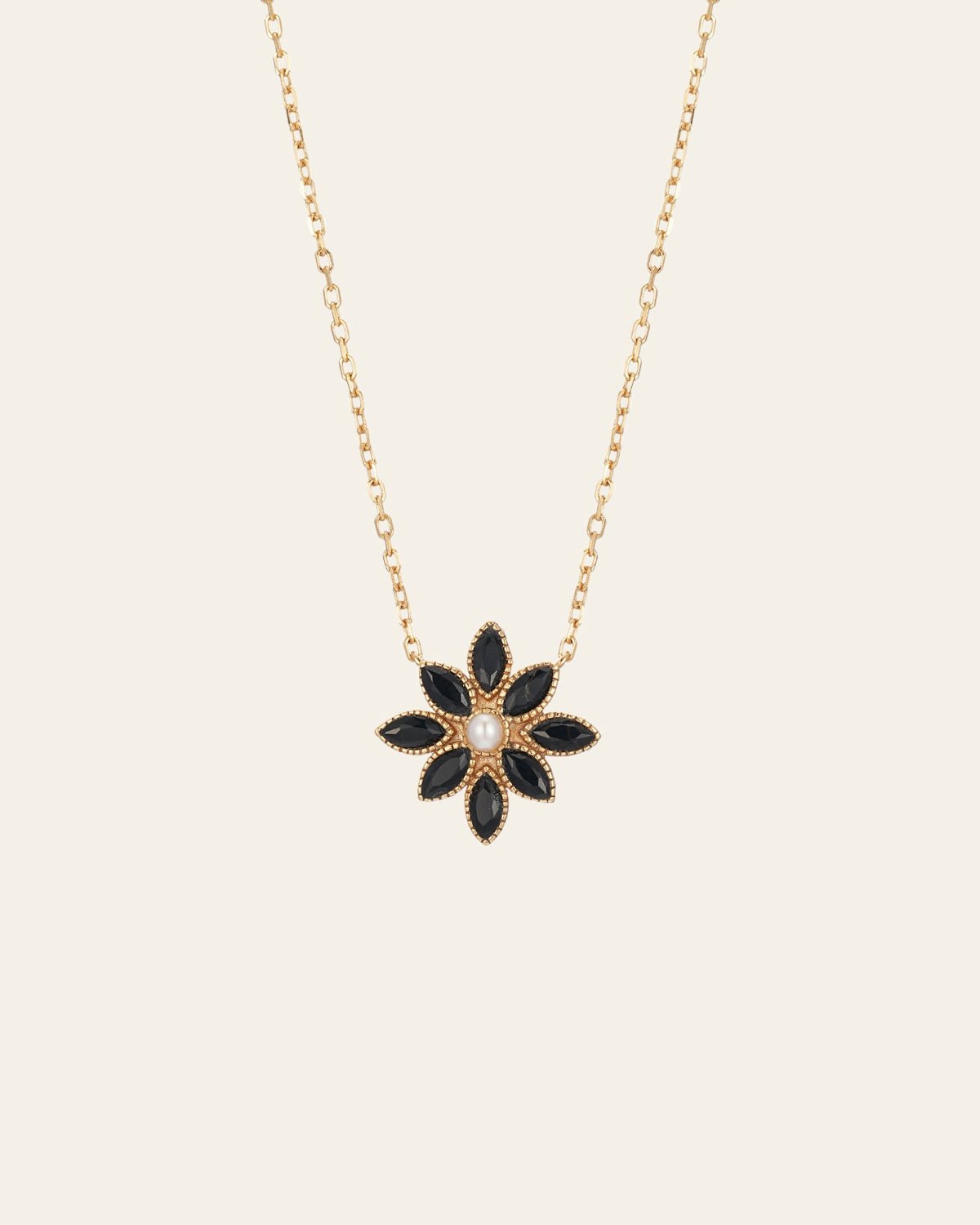 The CAMELLIA Cámellia Black Onyx Pearl Necklace features a gold chain with a floral pendant, highlighting six petal-shaped black onyx stones encircling a central pearl-like bead.