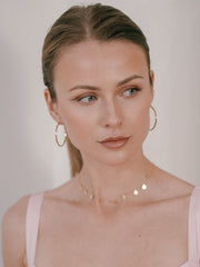 The woman, with her light brown hair tied back, wears CAMELLIA's 40mm Mother Hoops and a delicate, nickel-free hypoallergenic necklace. Gazing to the side with a neutral expression, she dons a pale pink top that complements her understated elegance.
