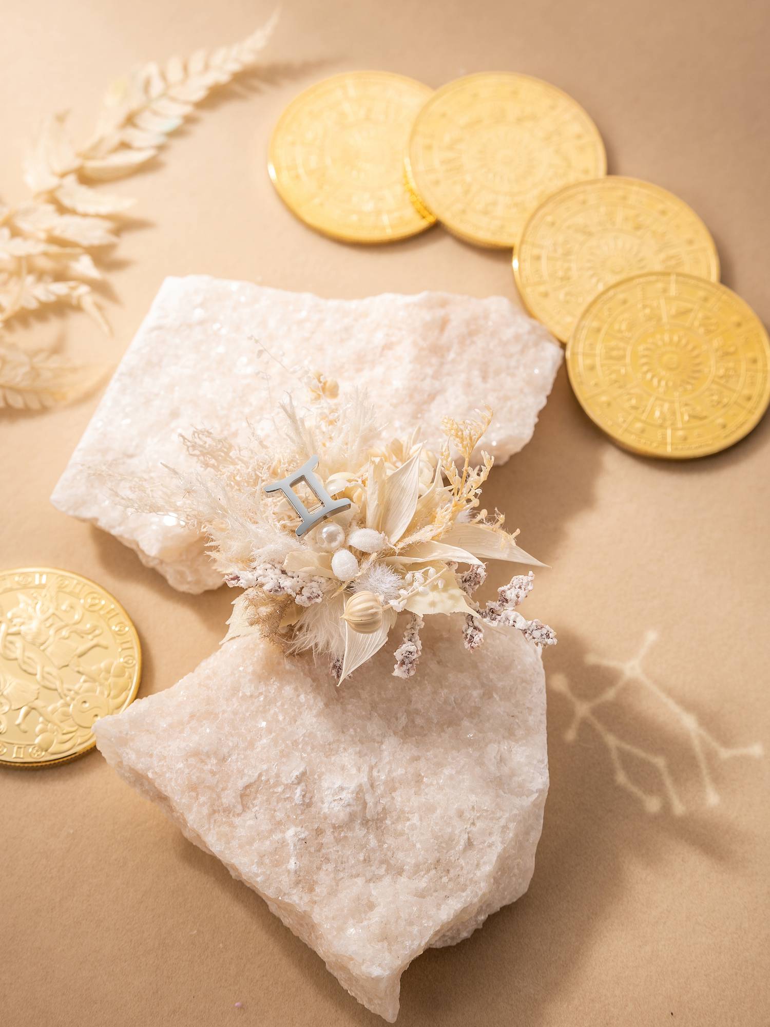 A small floral arrangement with CAMELLIA's Gemini Flower Ring, featuring a custom blue "H," rests on white stones and is surrounded by gold coins. Handcrafted in New York for a unique touch.
