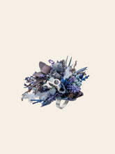 The Capricorn Flower Ring by CAMELLIA features a handcrafted silver design with dried purple, blue, and gray flowers, leaves, and branches. This adjustable piece is uniquely set against a plain background.