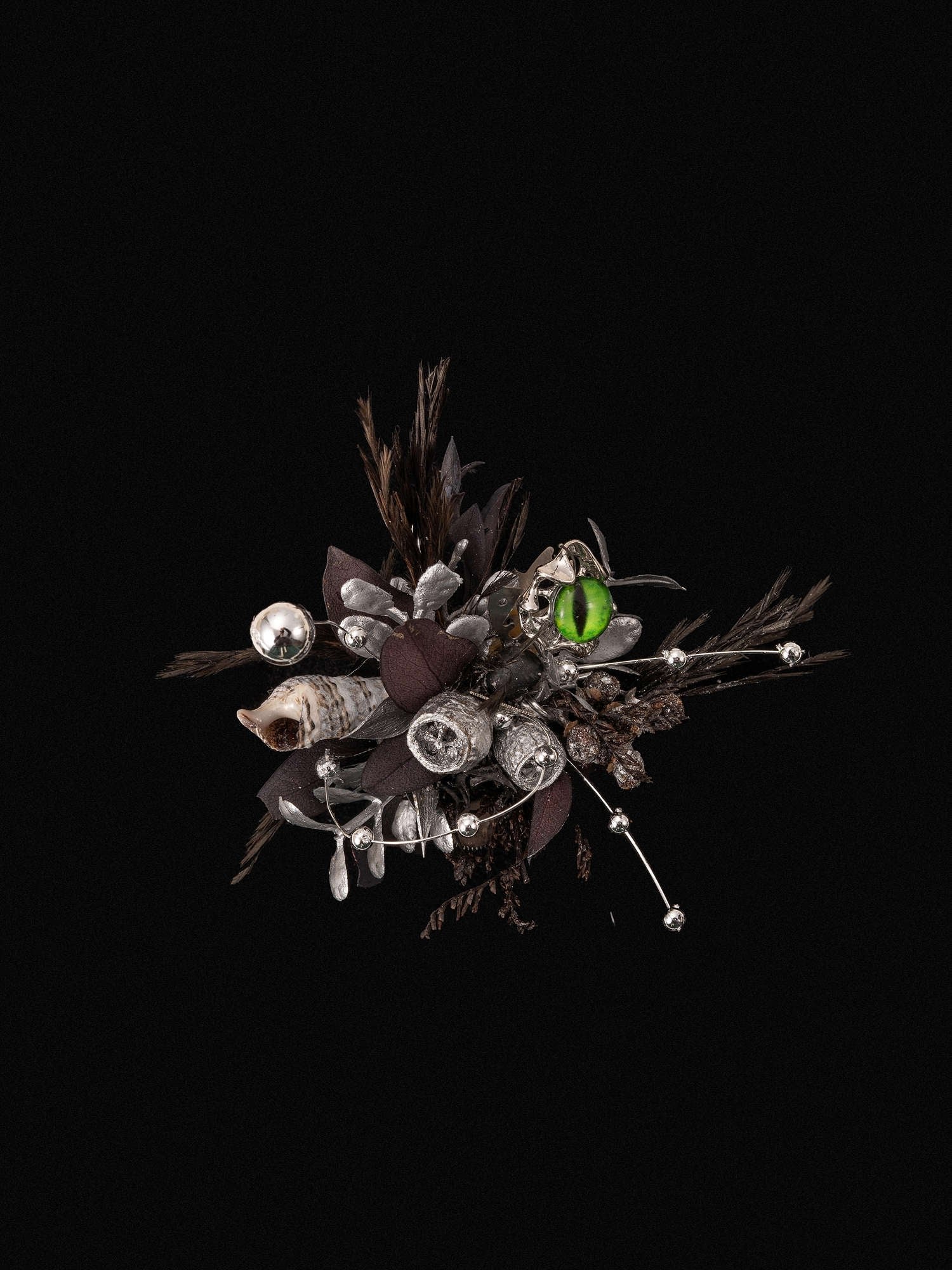 The Devil's Eye Flower Ring by CAMELLIA, featuring a detailed design of dried leaves, twigs, silver ornaments, a shell, and a bright green bead against a black background, captures the essence of handcrafted artistry in New York.