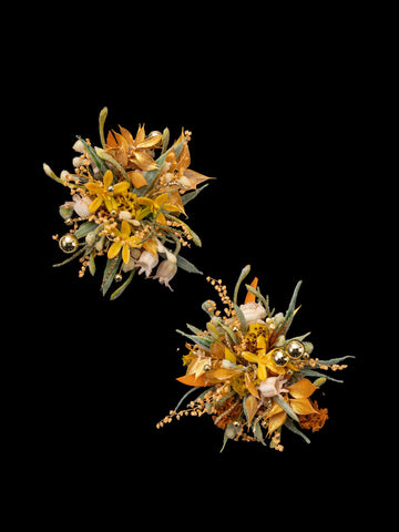 The CAMELLIA Love In The Sunset Wind Wedding Flower Rings feature two handcrafted arrangements with orange, yellow, and green elements, small gold spheres on a black background, and delicate dried flowers for added charm. The CAMELLIA Love In The Sunset Wind Wedding Flower Rings feature two handcrafted arrangements with orange, yellow, and green elements, small gold spheres on a black background, and delicate dried flowers for added charm.