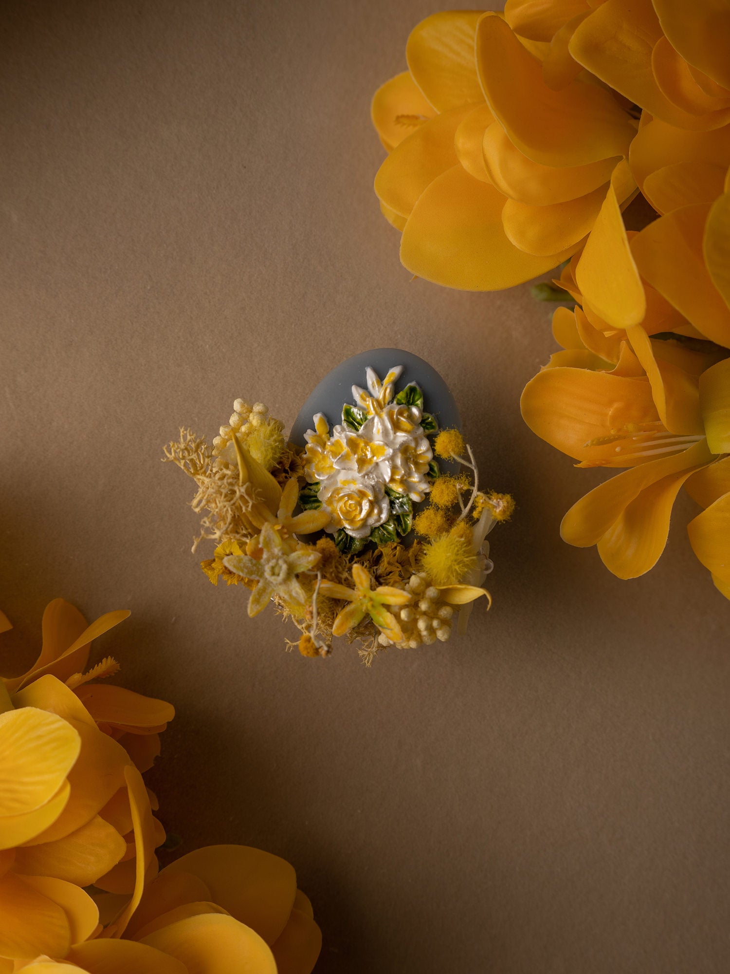 The Freesia Birth Flower Ring by CAMELLIA features a decorative blue egg with yellow and white floral designs nestled among vibrant orange flowers on a brown background, creating an elegant blend of color and nature's timeless allure.