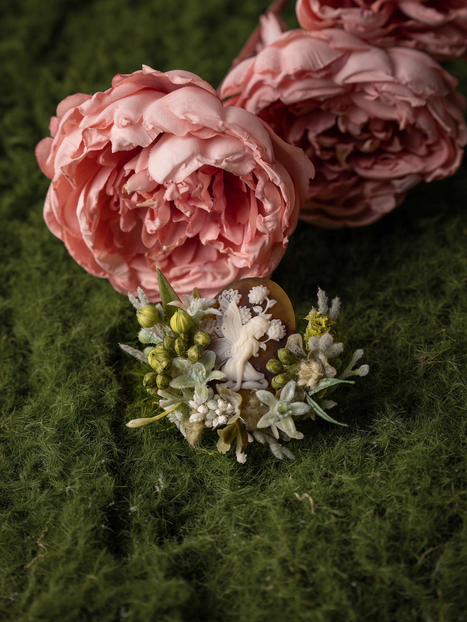 Delicately arranged pink roses on a green textured surface are enhanced by a display of dried preserved flowers and foliage. Adding an elegant touch is the handcrafted Peony Birth Flower Ring by CAMELLIA, offering a serene and sophisticated ambiance.
