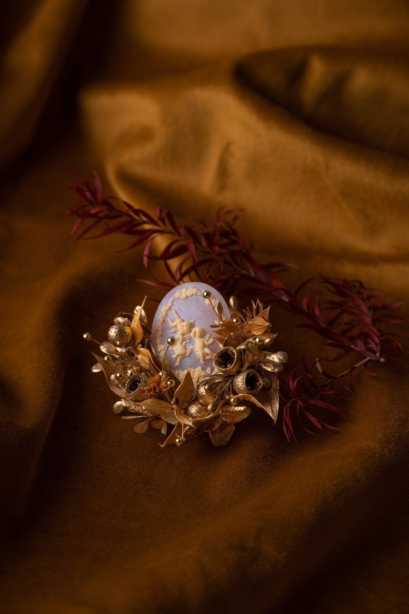 The Melaleuca Bracteata Birth Flower Ring by CAMELLIA is a handcrafted piece from New York, featuring a cameo center with gold floral accents on a brown fabric backdrop.