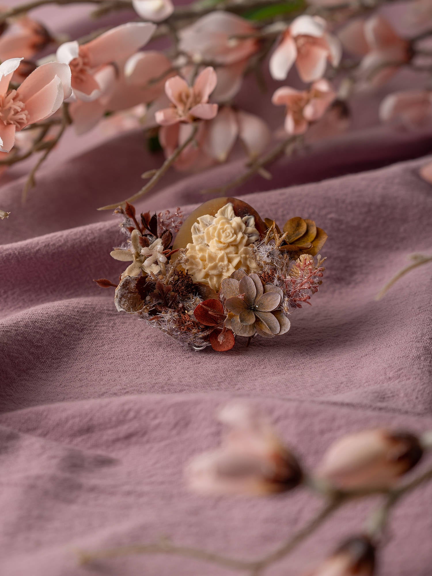 A decorative floral arrangement of dried flowers on soft pink fabric is surrounded by pink flower branches, enhanced by CAMELLIA's Magnolia Birth Flower Ring for a handcrafted jewelry charm.