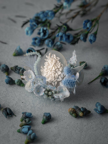 A decorative white egg with floral carvings is encircled by blue and white blooms on a textured gray surface, reminiscent of the elegance of CAMELLIA's Winteersweet Birth Flower Ring. Blue flower sprigs enhance this artistic display. A decorative white egg with floral carvings is encircled by blue and white blooms on a textured gray surface, reminiscent of the elegance of CAMELLIA's Winteersweet Birth Flower Ring. Blue flower sprigs enhance this artistic display.