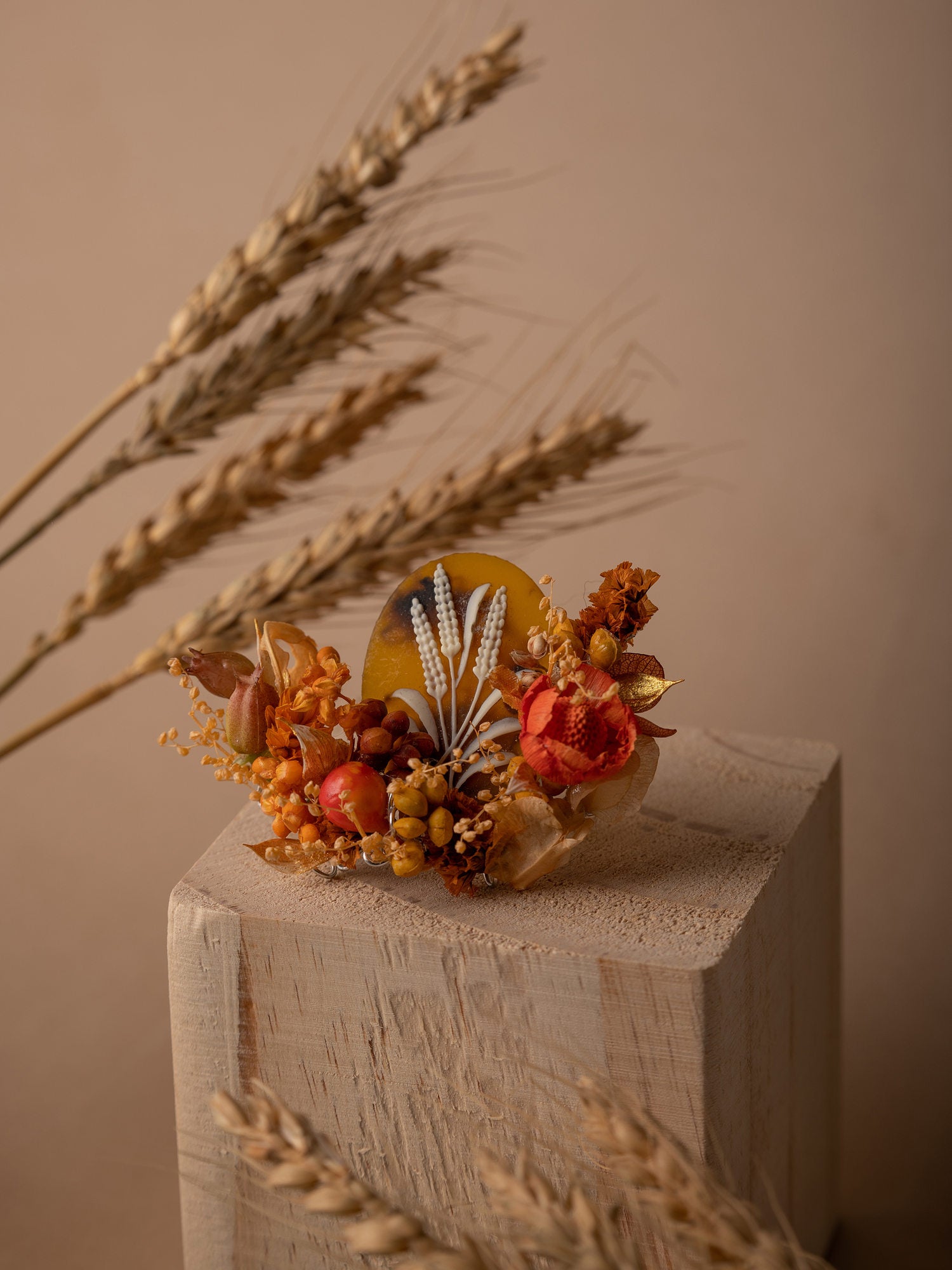 A Wheat Birth Flower Ring by CAMELLIA is showcased as a dried floral arrangement with wheat stalks, orange flowers, and a yellow accent on a wooden block against a beige background, exuding elegance with its New York artisanal craftsmanship.