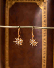 The Stella Radiance Opal Dangle Earrings by CAMELLIA feature gold star shapes with pearl centers and diamond accents elegantly hanging on a thin rod. The opulent design is highlighted against a brown and gold backdrop with hints of yellow gold enhancing their intricate beauty.