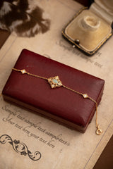 The Aurielle Opal Bracelet by CAMELLIA shines with diamond-shaped details and a central embellishment atop a closed red leather box. Nearby, a vintage necklace box gleams, its brilliance rivaled by the opal accents in the yellow gold design.