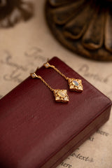CAMELLIA's Aurielle Opal Dangle Earrings, featuring gold chains and ornate square pendants, stylishly rest on a closed maroon book. These hypoallergenic beauties offer unmatched style and comfort for sensitive skin, with a brass object subtly visible in the blurred background.