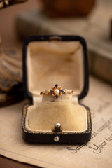 The CAMELLIA Chloe Pearl Tanzanite Jewelry Set, featuring a gold ring with a central pearl, tanzanite, and clear stones, rests elegantly in an open vintage jewelry box on soft fabric lining.