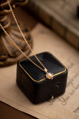 The Chloe Pearl Tanzanite Jewelry Set by CAMELLIA features a gold necklace with a floral pendant and tanzanite accents, resting on a black box atop a textured sheet, with a blurred background.