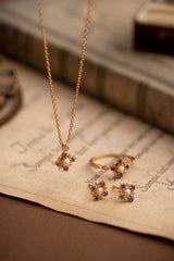 The Chloe Pearl Tanzanite Jewelry Set by CAMELLIA features a stunning yellow gold necklace, ring, and earrings with pearl and tanzanite accents, elegantly showcased on vintage paper with a softly blurred book in the background.