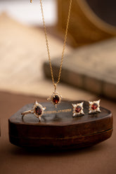 The Claire Garnet Diamond Jewelry Set by CAMELLIA, featuring a necklace, ring, and earrings adorned with vibrant garnet gemstones, is elegantly displayed on a brown leather box.