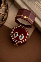 A pair of gold earrings from the Louisa Sapphire Jewelry Set by CAMELLIA, crafted with 14K yellow gold vermeil and featuring rectangular blue sapphires encircled by shimmering diamonds, is beautifully displayed in an open red velvet box on a brown surface.