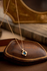 The Louisa Sapphire Jewelry Set by CAMELLIA features a 14K yellow gold vermeil necklace with a stunning blue gemstone pendant, elegantly displayed on a wooden surface with books in the background.