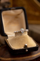 The CAMELLIA Starlight Opal Jewelry Set features a handcrafted teardrop-shaped opal ring with diamond accents set in sterling silver, elegantly displayed in a vintage velvet-lined box on a wooden surface.
