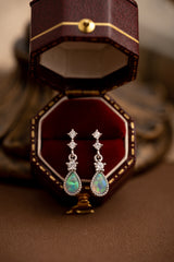 A pair of handcrafted teardrop-shaped opal and diamond earrings from the Starlight Opal Jewelry Set by CAMELLIA rests in an open red velvet box, set against a soft-focus background.
