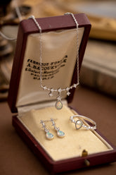 The CAMELLIA Starlight Opal Jewelry Set includes a handcrafted necklace, earrings, and ring in an open brown box. These Sterling Silver pieces feature exquisite opal gemstones with diamond-like accents.