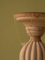 The CAMELLIA 555 Angel Number Choker with Peridot is displayed draped over a textured beige ceramic vase, set against an olive green backdrop.