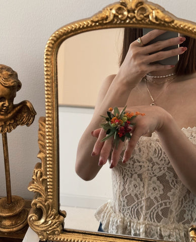 A person takes a mirror selfie, wearing a lace top and holding flowers. The ornate mirror and decorative angel statue are visible, while CAMELLIA's Customized Basic Flower Ring, handcrafted in New York, subtly enhances their elegant look. A person takes a mirror selfie, wearing a lace top and holding flowers. The ornate mirror and decorative angel statue are visible, while CAMELLIA's Customized Basic Flower Ring, handcrafted in New York, subtly enhances their elegant look.