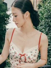 A person with dark hair tied back, in a light dress with red straps and cherry patterns, stands outdoors near greenery. They're wearing CAMELLIA's Elsa Rose Pearl Earrings, which catch the sunlight, adding a subtle touch of elegance to the scene.