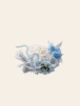 The Winteersweet Birth Flower Ring by CAMELLIA showcases a delicate arrangement of dried preserved white flowers and blue accents with beads and textured foliage on a simple beige background.