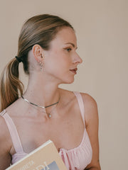 A woman with a ponytail and earrings holds a book while wearing a pink sleeveless top and the elegant Elsa Peony Sapphire Necklace from CAMELLIA, crafted in 14K Yellow Gold Vermeil.
