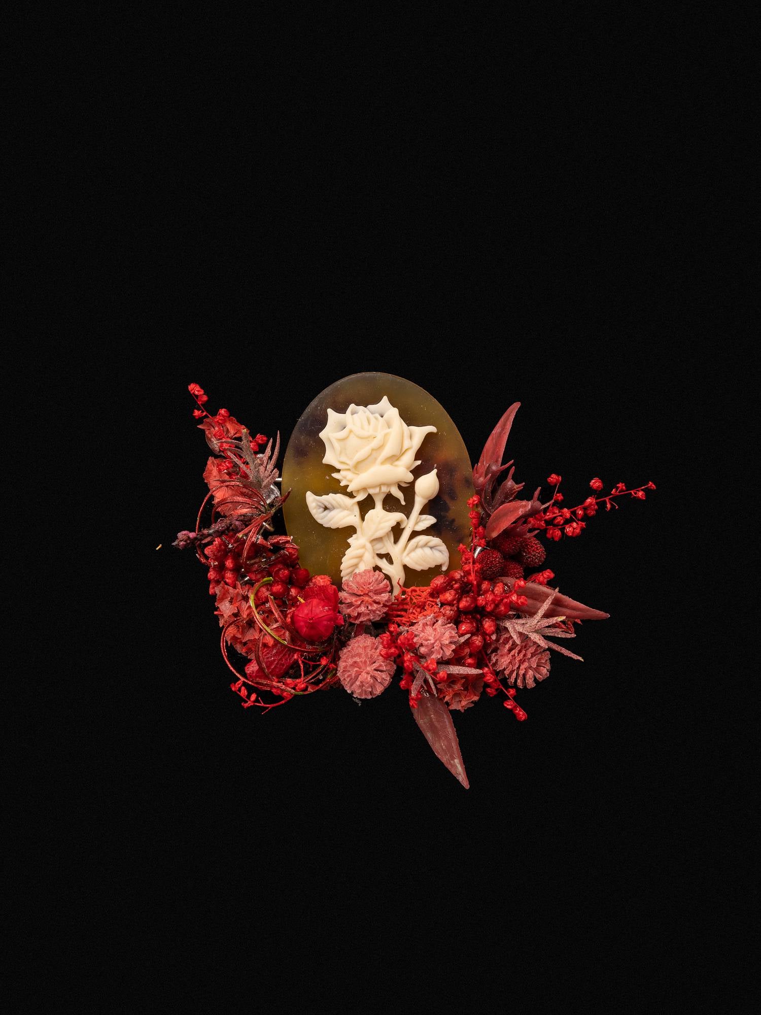 The Rose Birth Flower Ring by CAMELLIA features a detailed white rose on a dark oval, surrounded by red and pink dried flowers. Handcrafted in New York, it offers an adjustable size for a perfect fit.