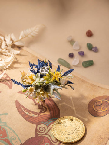 A small bouquet of artificial flowers and leaves rests on a decorative surface with a golden coin and tiny gemstones in the background, resembling the CAMELLIA Cancer Flower Ring handcrafted in New York. A small bouquet of artificial flowers and leaves rests on a decorative surface with a golden coin and tiny gemstones in the background, resembling the CAMELLIA Cancer Flower Ring handcrafted in New York.