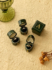 CAMELLIA's Customized Vintage Ring Boxes, adorned with assorted rings, are beautifully displayed on a textured surface alongside dried flowers and a green glass cup.
