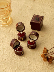 Three open CAMELLIA Customized Vintage Ring Boxes, each with a differently styled ring, sit on a textured surface next to a glass and dried flowers. In the background is a closed CAMELLIA box featuring an "M" logo.