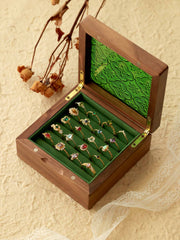 An open wooden box reveals a collection of ornate CAMELLIA rings, including the stunning Mystery Gemstone Ring in yellow gold and sterling silver, all displayed on a green velvet lining resting on a textured surface with dried flowers and lace for an elegant scene.